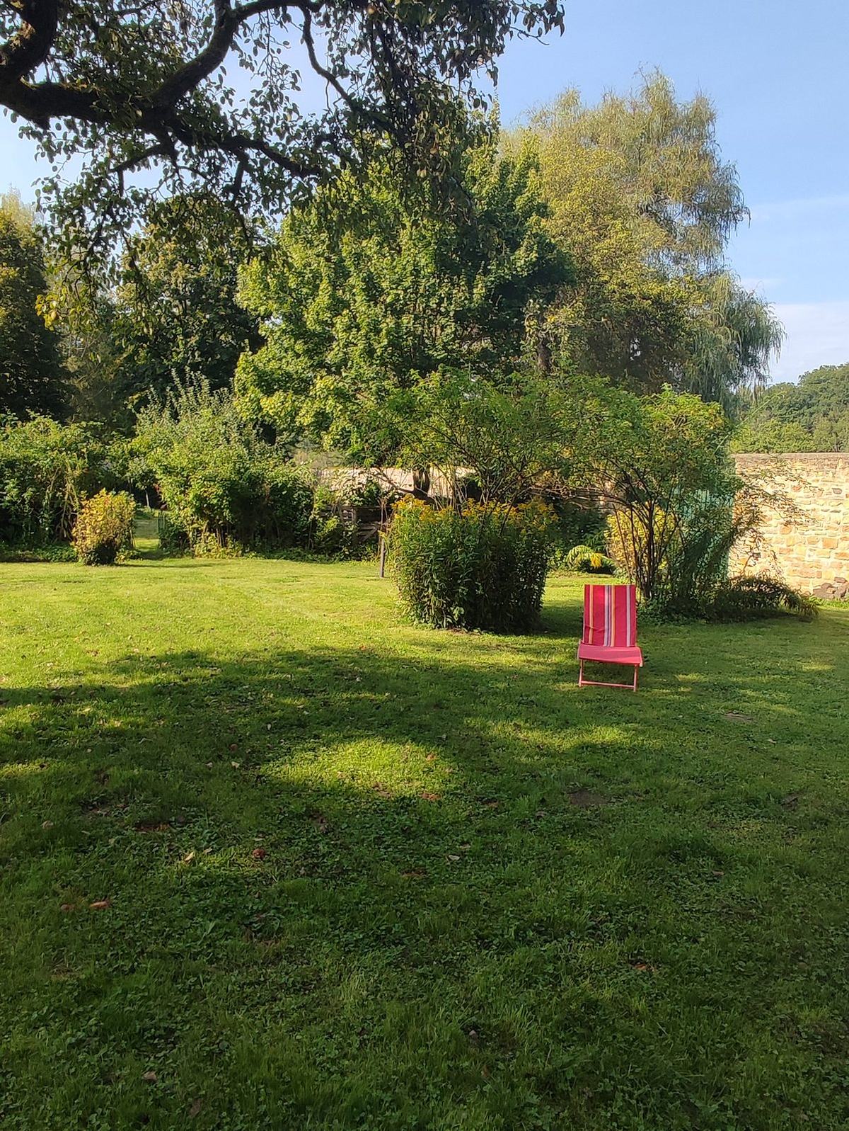 Grande pelouse tondue avec un fauteuil rouge posé au milieu, massifs fleuris et mur de pierre au fond.