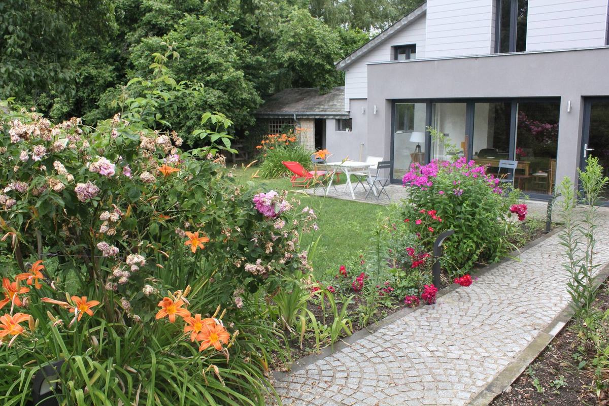Le jardin fleuri du gîte en été : hémérocalles orange et rosiers au premier plan, allée pavée menant à la terrasse et aux baies vitrées de la maison.