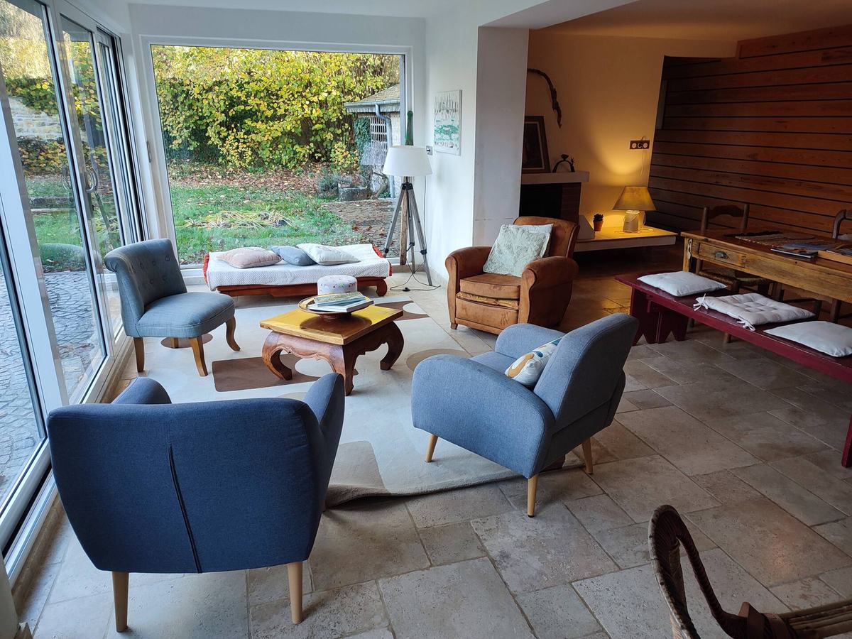 Le salon : fauteuils bleus autour d’une table basse en bois, sol en travertin, baies vitrées ouvrant sur le jardin.