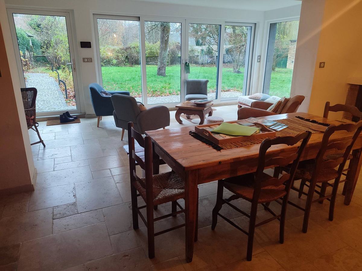 La véranda et la salle à manger : table en bois dressée, chaises anciennes, baies vitrées d’angle sur le jardin.